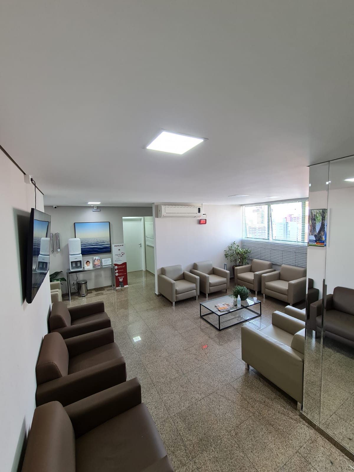 Sala de espera da Clínica PrimeCor com poltronas bege e marrom, mesa de centro de vidro, piso de granito e um quadro de mar ao fundo